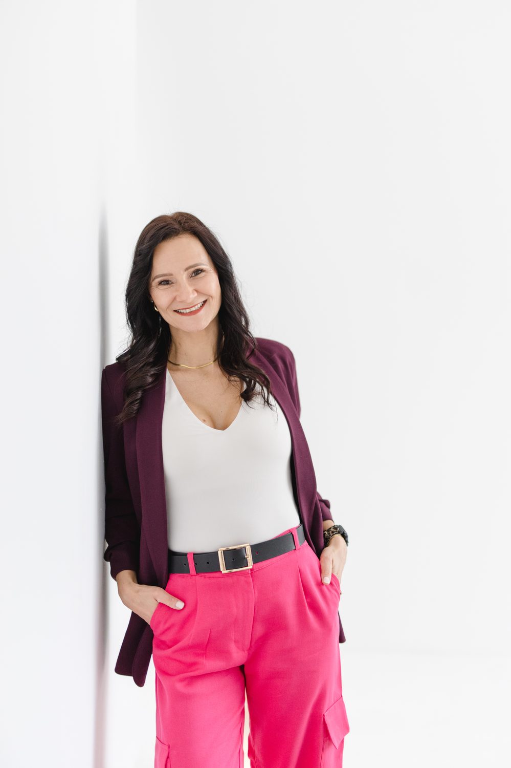 Karolina Dudek lehnt lächelnd an einer weißen Wand – in Beeren-Blazer und pinker Hose
